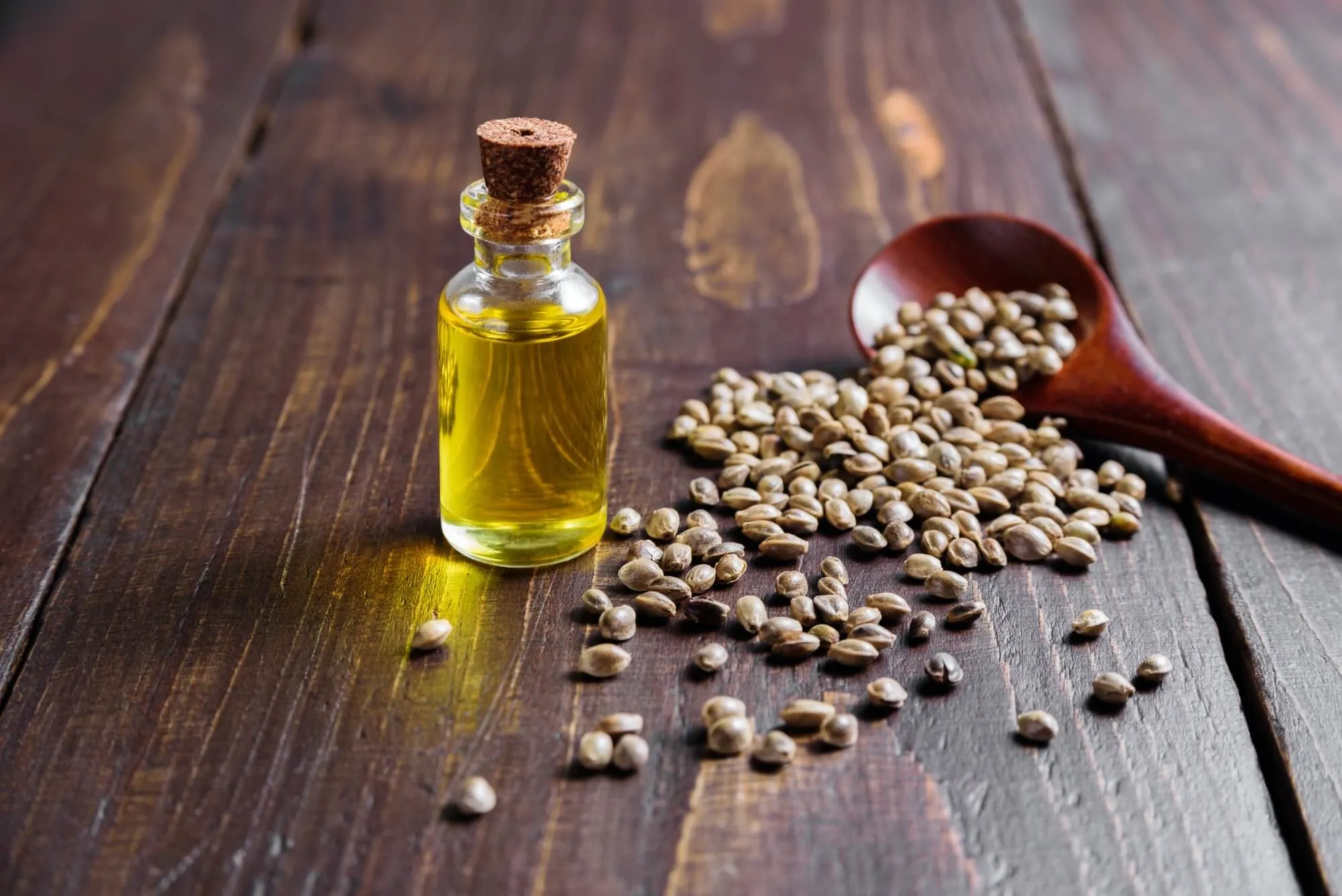 A bottle of hemp seed oil beside a spoon of hemp seeds on a wooden table