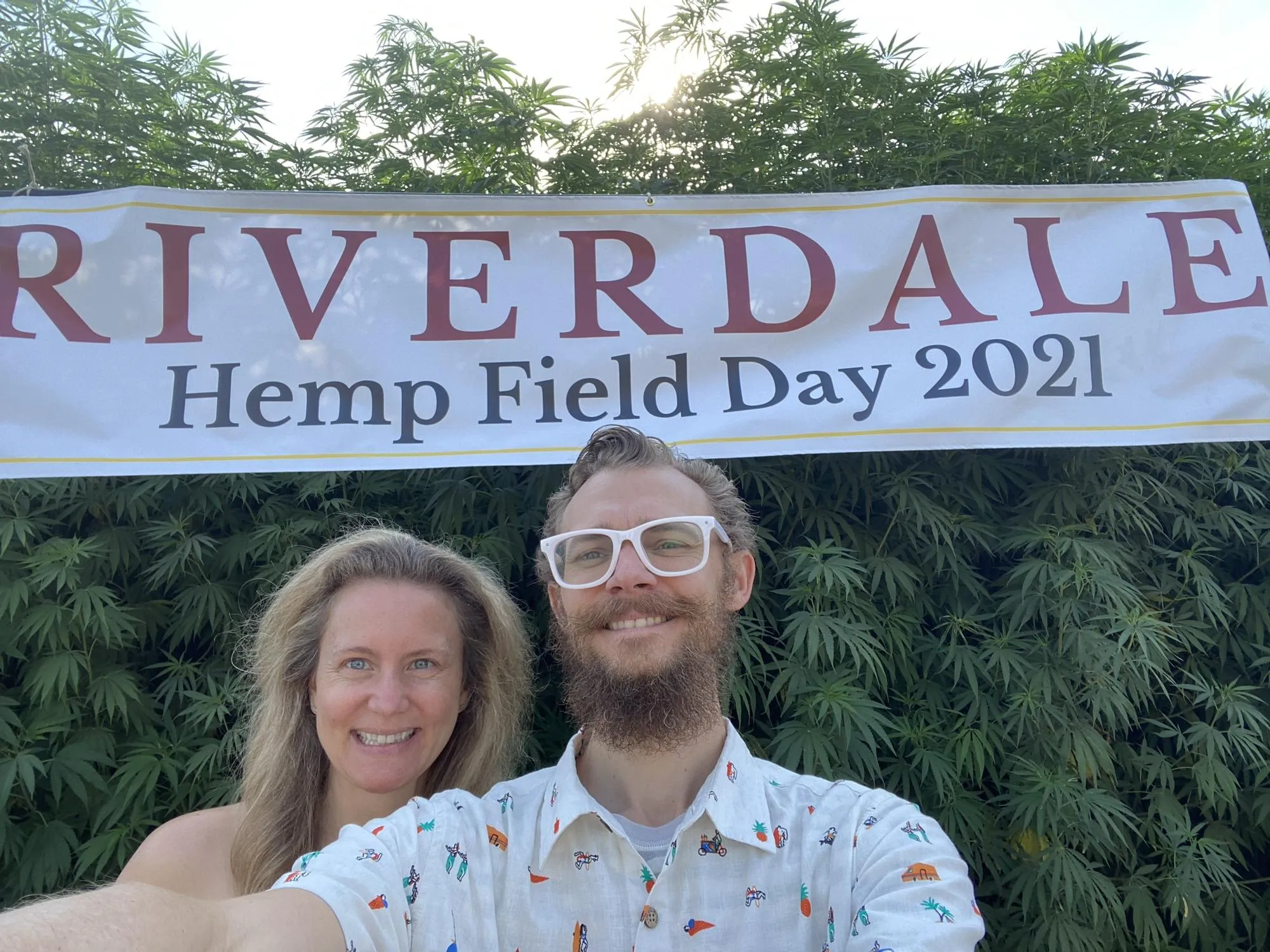 Tyler Hemp and Dodee in front of a wall of hemp plants under a banner reading Riverdale Hemp Field Day 2021